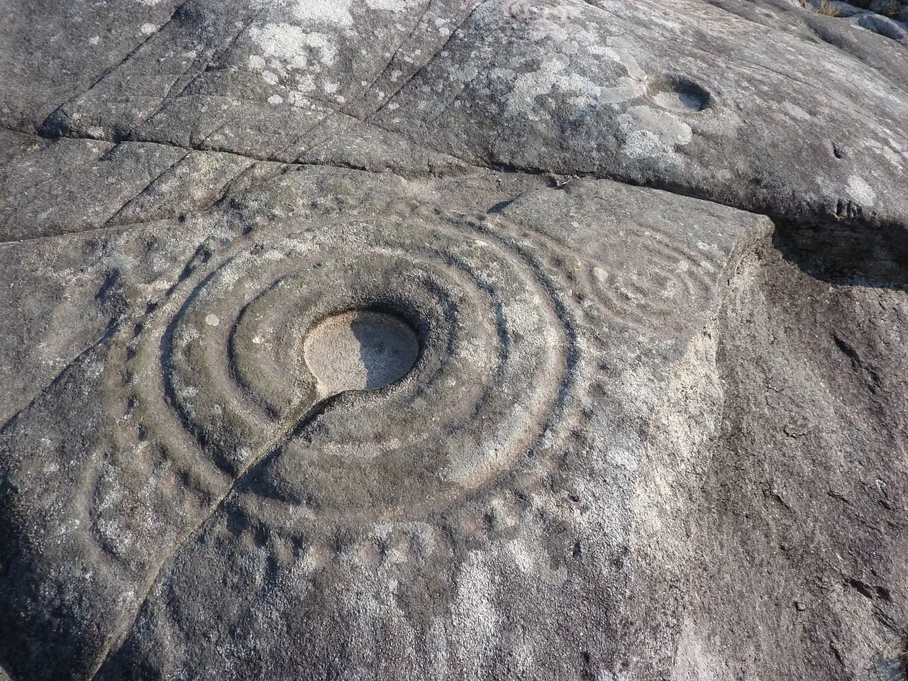 Petroglifo de círculos concéntricos con canal central en Laxe das Rodas, Galicia, obsérvense las pequeñas cazoletas que rodean la figura como en Marruecos. Son especialmente abundantes en toda la cornisa atlantica europea / Wikipedia